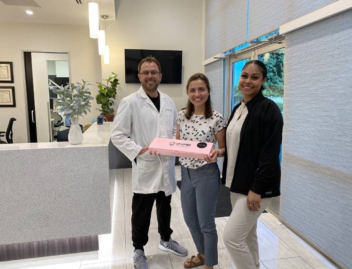 Doctor Rodda posing with two young women bringing pastry box into dental office